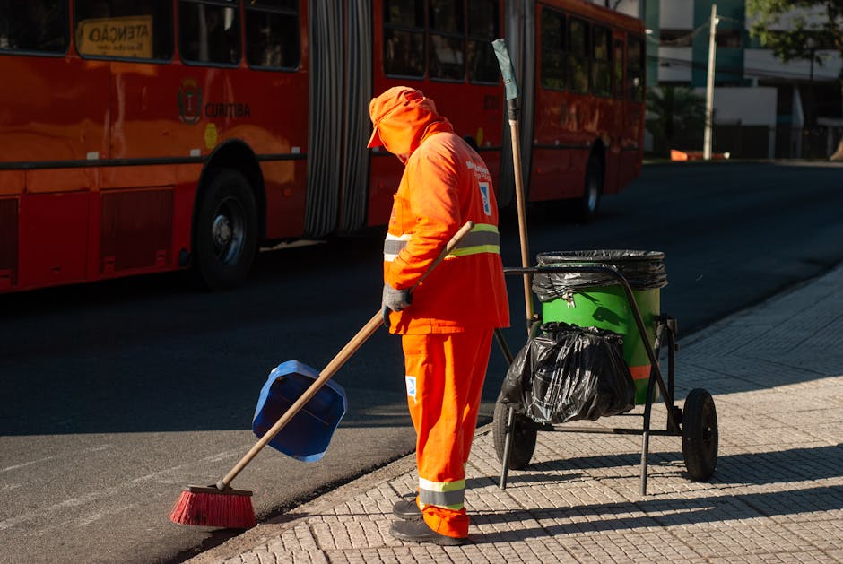 A street scene in Bromley with several pedestrians, including a sanitation worker in a green uniform and orange safety vest, emptying a large black wheeled trash bin, which is on a paved brick walkway