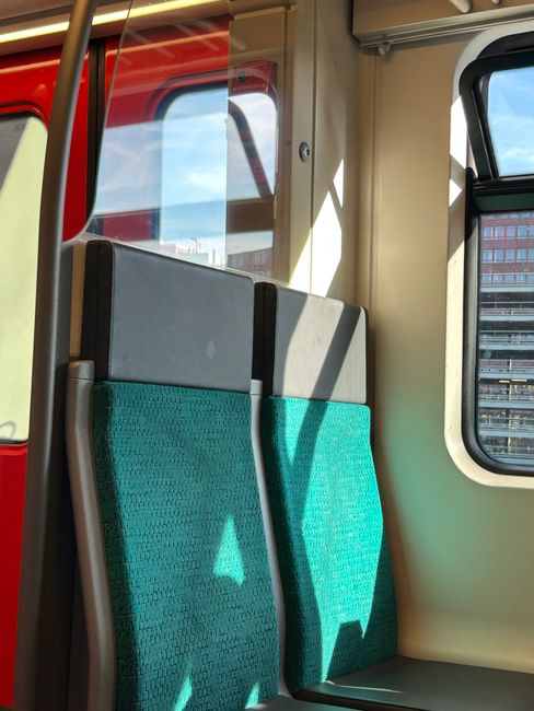 Interior view of a clean, empty train carriage on the London Underground, with blue handrails, patterned fabric seats, and clear windows. The carriage is well-lit with natural and artificial light, an