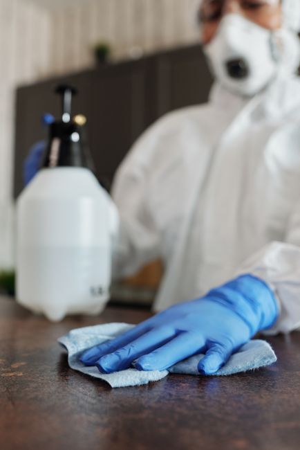 A person dressed in a white protective suit with blue gloves is holding a spray gun connected to a 1.5-liter container, performing surface sanitisation on a kitchen countertop. The scene shows a moder