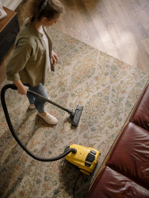 A person operating a yellow portable carpet cleaning machine on a patterned area rug, with the individual's hands visible pressing buttons. The machine has a built-in vent for air circulation and a wa